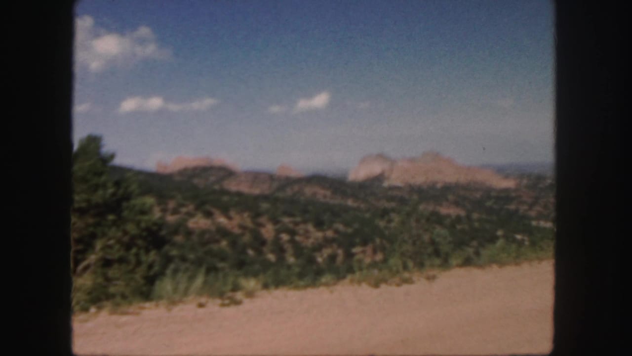 1958 Colorado 8mm frame showing a dirt road edge, green shrubs, rolling hills, and distant reddish rock formations beneath scattered clouds.