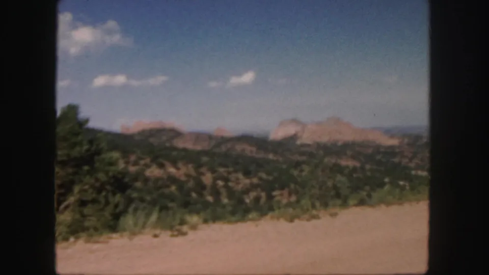 1958 Colorado 8mm frame showing a dirt road edge, green shrubs, rolling hills, and distant reddish rock formations beneath scattered clouds.