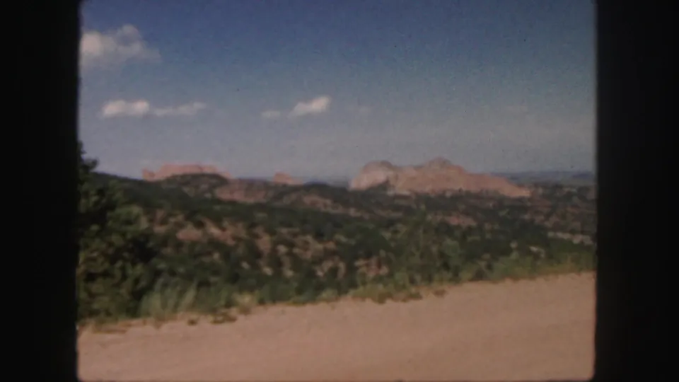 1958 Colorado home-movie frame with a dusty roadside foreground, wooded scrub hills, distant red rock ridges, and scattered white clouds.