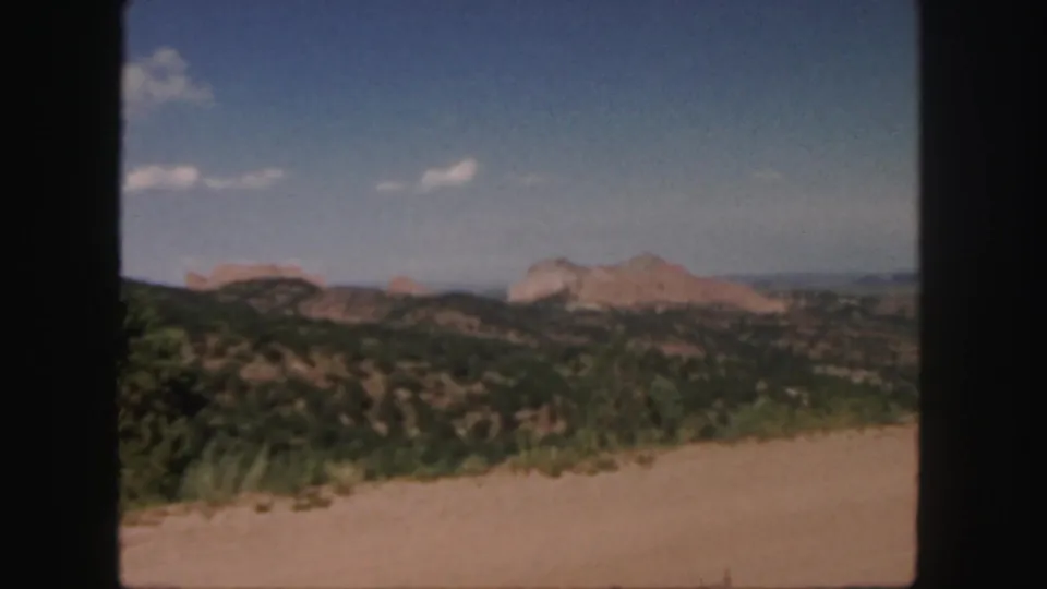 1958 Colorado frame looking over a pale dirt road toward scrub-covered terrain and reddish rock formations beneath a clear blue sky.