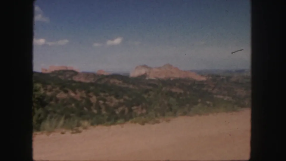 1958 Colorado 8mm frame showing a dusty road edge, dark green shrubs, distant red rock hills, blue sky, and a small film mark near the right sky.