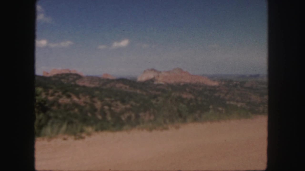 1958 Colorado 8mm frame showing tan foreground, green scrub hills, and distant red rock formations beneath scattered clouds.