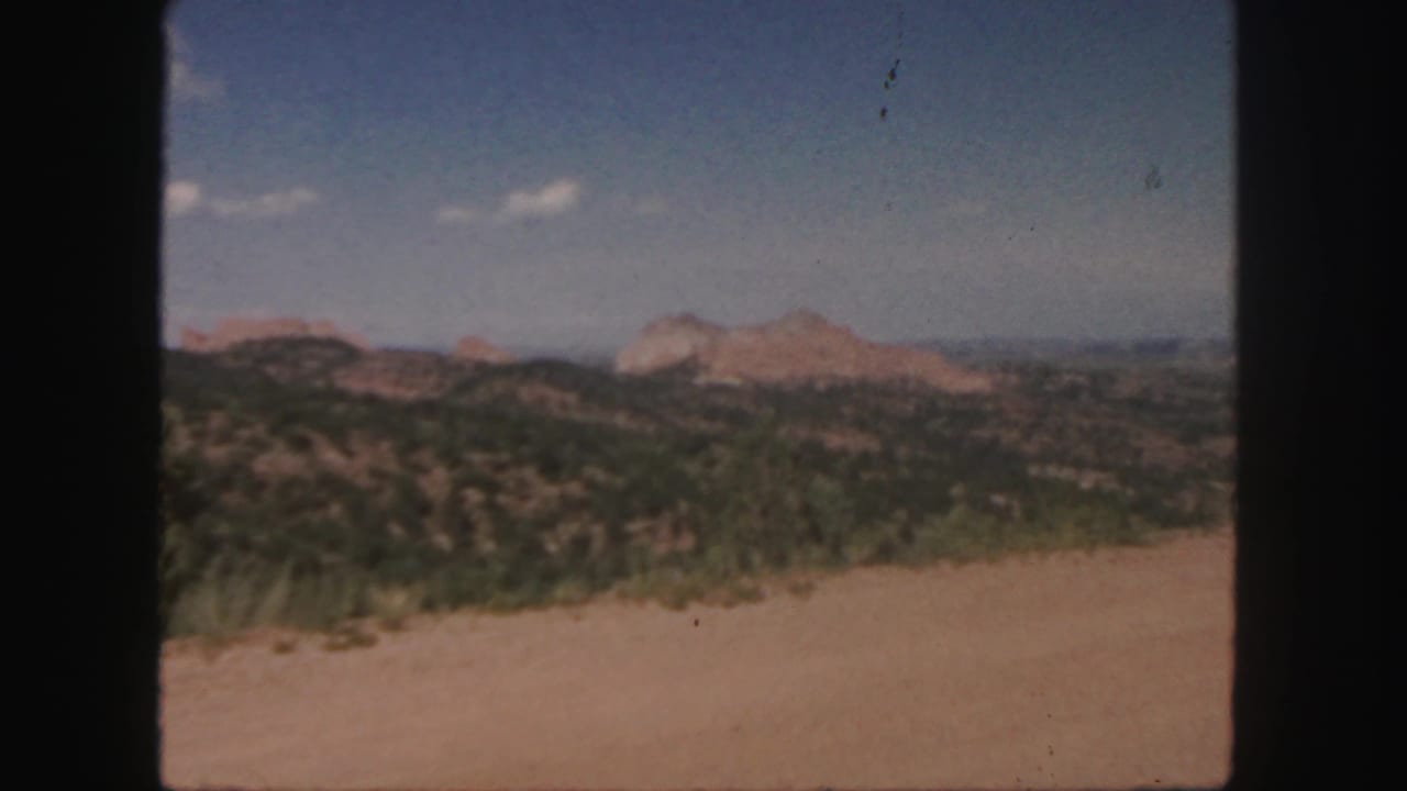 Shrubland hills lead toward pale red rock ridges below a clear sky.