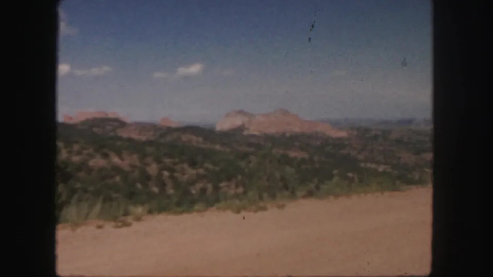 Vintage 8mm Colorado landscape frame with rolling green terrain, distant reddish rock outcrops, and visible film gate edges.