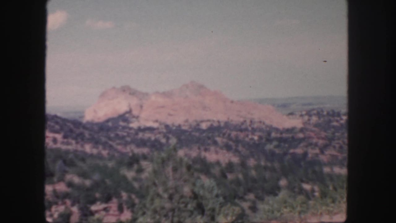 1958 Colorado 8mm frame showing rugged reddish mountains beyond dry scrub and low trees under a hazy blue sky.