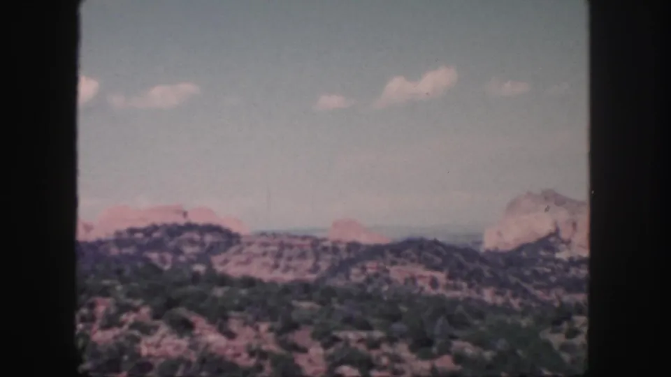 1958 Colorado 8mm frame showing a wider high-desert panorama with reddish rock outcrops, low hills, and a distant mountain edge.