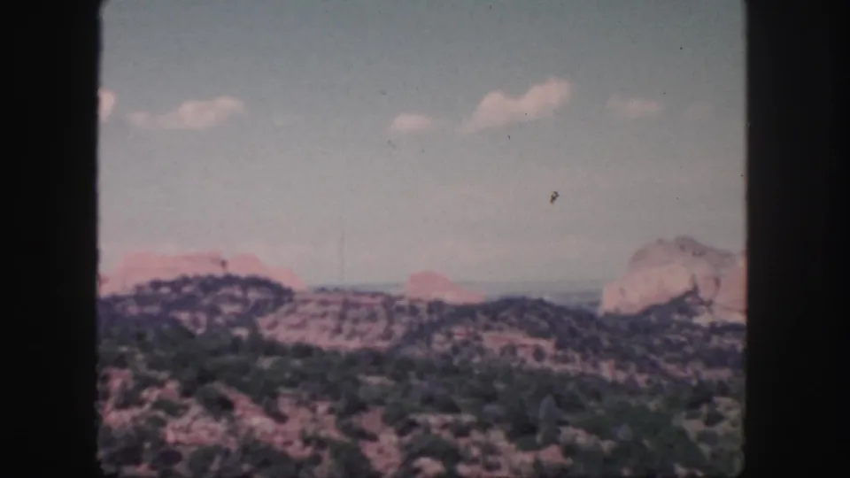 1958 Colorado home-movie frame showing arid red rock hills, dark shrubland, and a pale sky with scattered clouds.