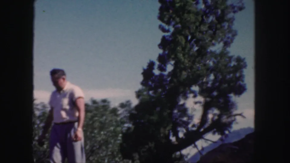 In 1958 Colorado 8mm footage, an adult man stands outdoors near orchard trees with distant hills behind him.