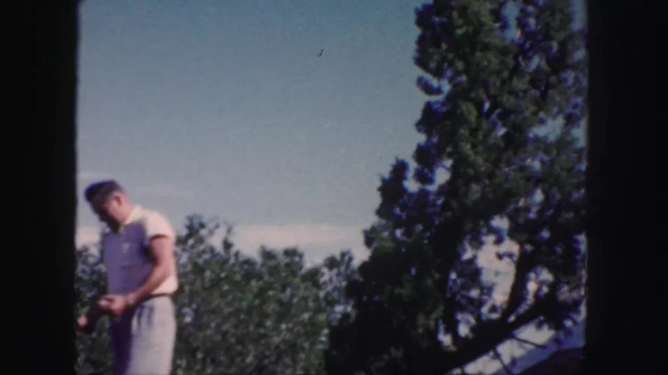 The man looks down at his hands in a Colorado orchard scene, framed by trees and blue sky on 8mm film.