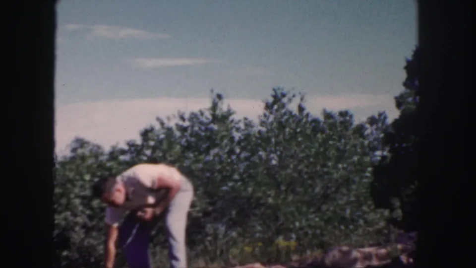 The man bends low in the orchard, reaching toward the ground for fallen fruit in 1958 Colorado home-movie footage.