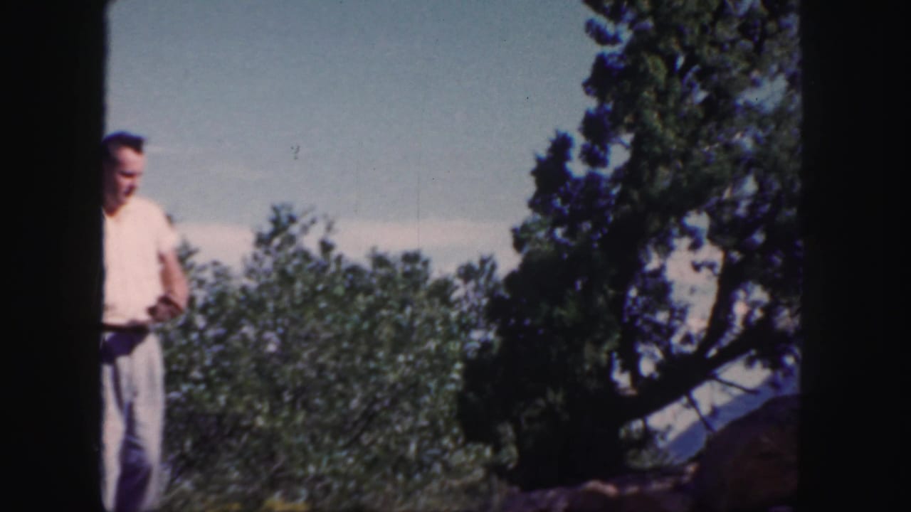 In 1958 Colorado 8mm footage, a man stands near evergreens and rocks with distant mountains under a clear sky.