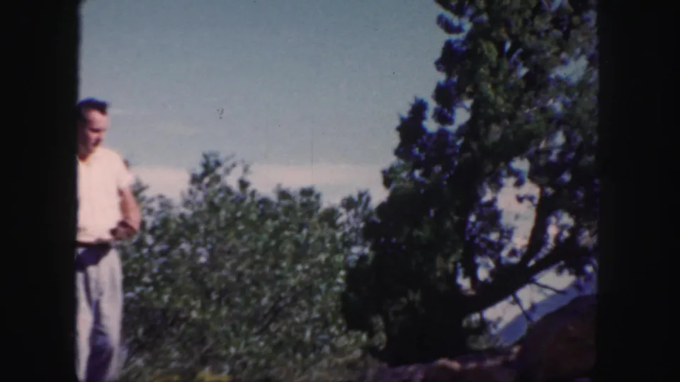 In 1958 Colorado 8mm footage, a man stands near evergreens and rocks with distant mountains under a clear sky.