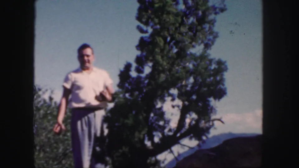 The man approaches the camera on a rocky Colorado ridge, with evergreens and distant peaks visible in the 1958 home movie frame.