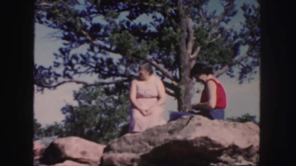 Two women sit on a boulder beneath an evergreen tree in the Colorado mountain landscape, one in a pale dress and one in a red top.