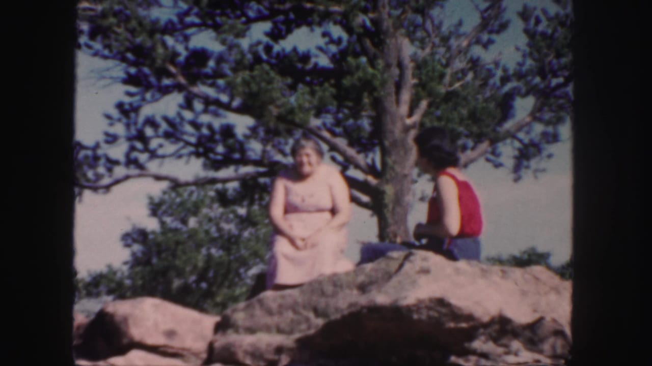 1958 Colorado home-movie frame with two women relaxing on a rock under a pine tree.