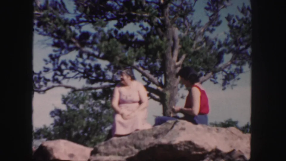 1958 Colorado home-movie frame of two women resting on a rocky ledge under pine branches.