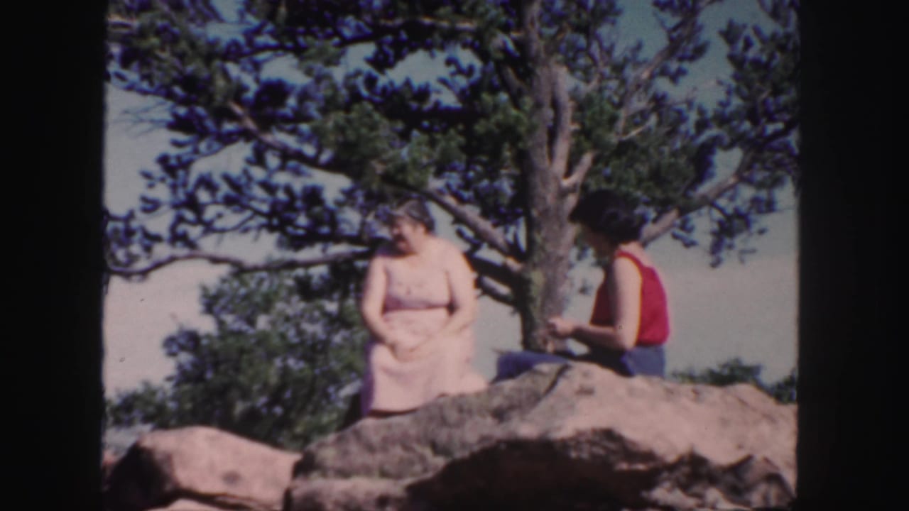 1958 Colorado 8mm frame showing two women seated on a pale rock under pine branches, with rugged rocks and sky behind them.
