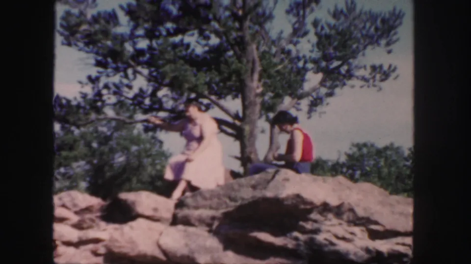 1958 Colorado 8mm frame showing one woman moving down from the rocks while another sits beside the pine tree.