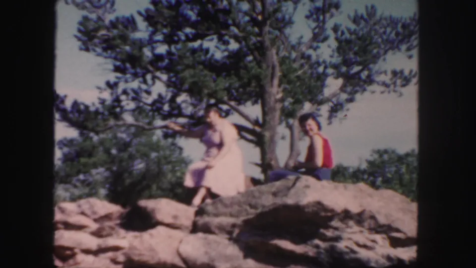 1958 Colorado home-movie still of two women seated among large rocks, with pine branches and foliage in the background.