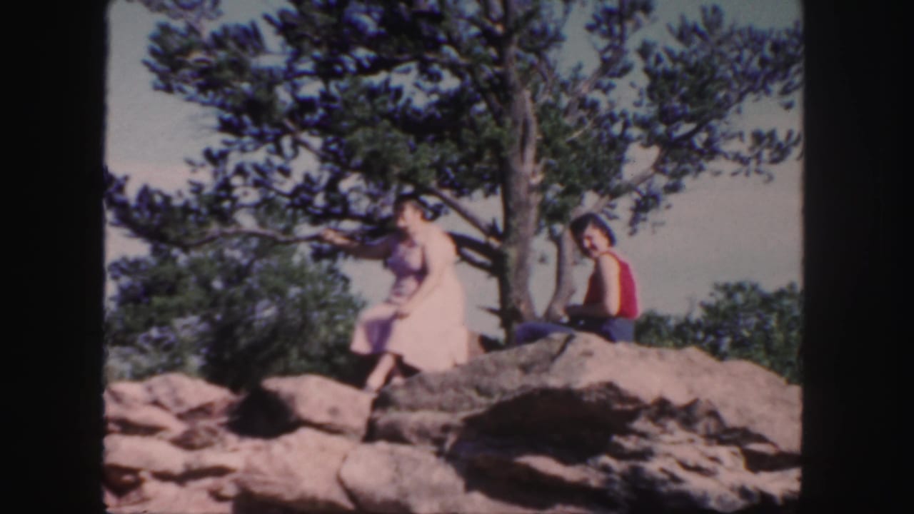 1958 Colorado 8mm frame showing a woman in a pink dress on a rocky outcrop near a tree, with another person in a red top beside her.