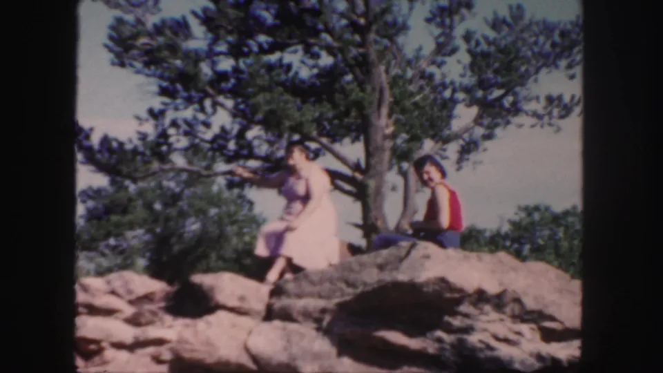 1958 Colorado 8mm frame showing a woman in a pink dress on a rocky outcrop near a tree, with another person in a red top beside her.