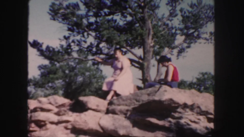 1958 Colorado 8mm frame showing two people seated on rocks under a tree, with the woman in pink turned toward the landscape.