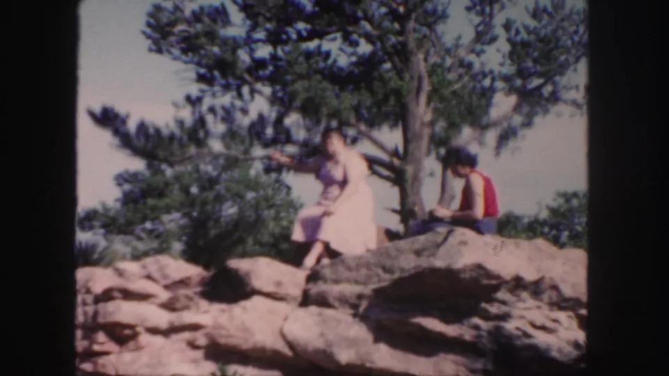 1958 Colorado home-movie frame of a woman in a pink dress sitting on sunlit rocks near a tree with another person beside her.