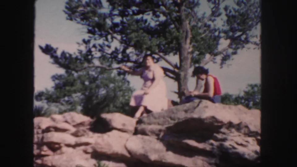 Two adults in 1958 Colorado home-movie footage sit on sunlit boulders beneath a pine tree, looking across the landscape.