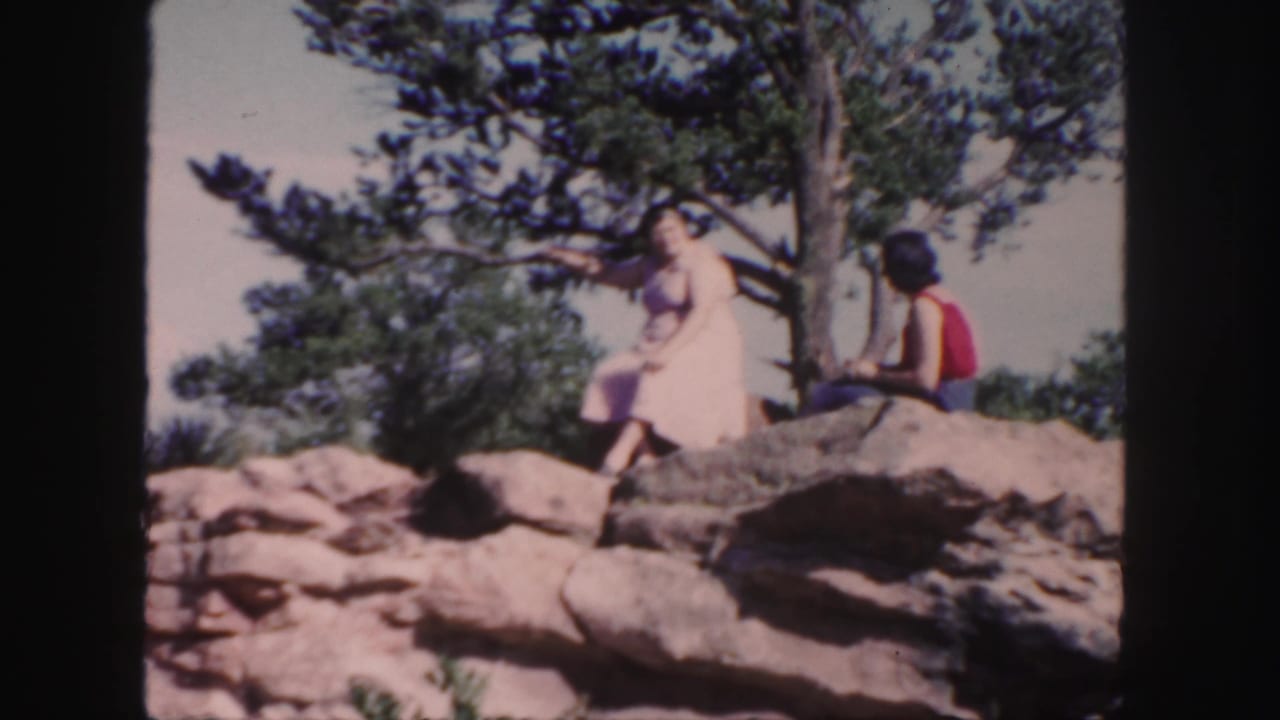 A woman in a light dress and a man in a red sleeveless top sit on a rocky Colorado overlook in 1958 home-movie footage.