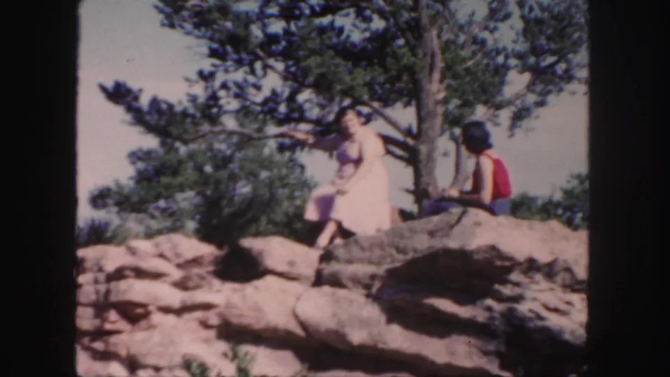 A woman in a light dress and a man in a red sleeveless top sit on a rocky Colorado overlook in 1958 home-movie footage.
