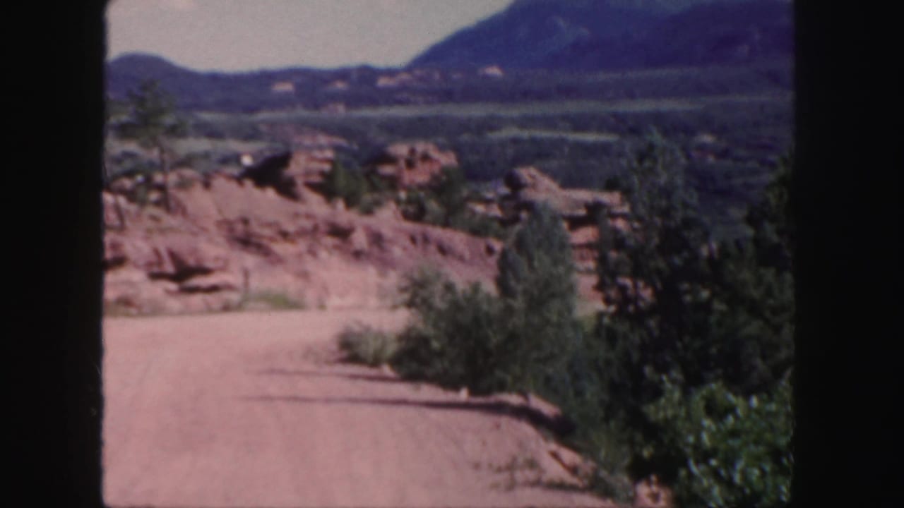Dusty road curving through red rock terrain with shrubs and distant mountains.