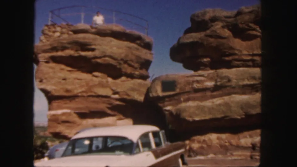A tourist stands behind a railing atop a red rock formation in Colorado, with a white 1950s car parked below.