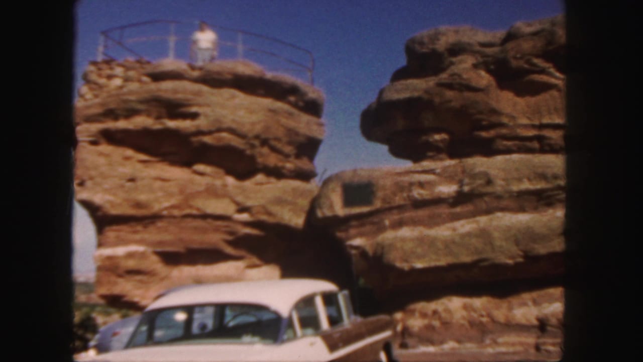 1958 Colorado home-movie frame showing a man on a sandstone overlook with a metal railing and a classic car below.