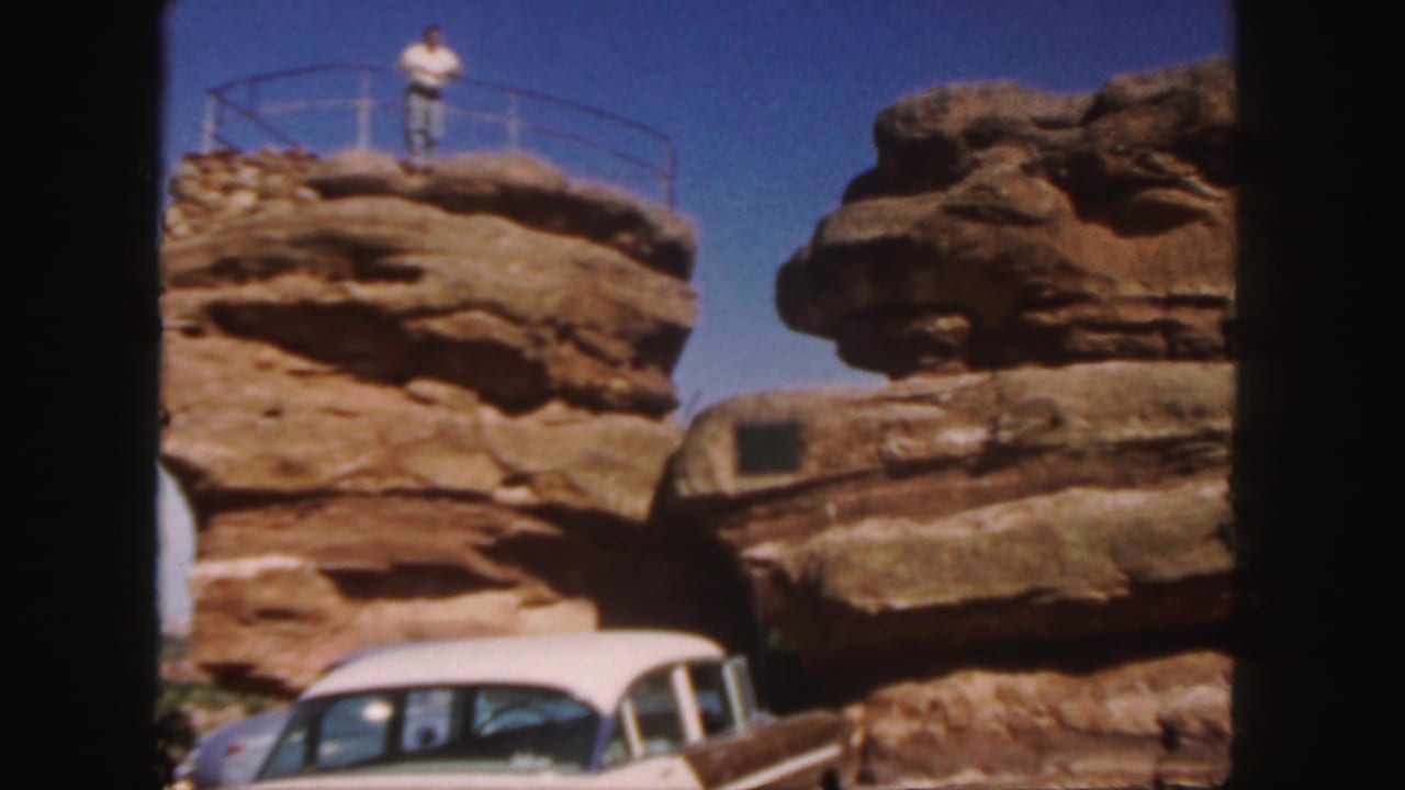 Man on a rock formation above a classic car and sandstone layers.
