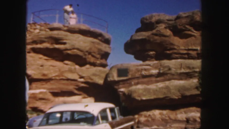 A man and woman stand together on the sandstone overlook above the parked car in this 1958 Colorado home-movie frame.