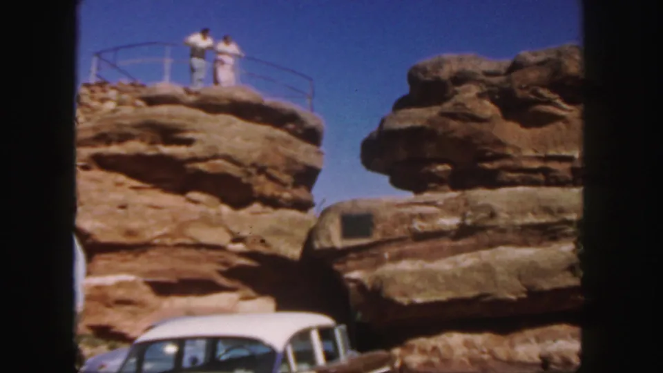 The couple poses on the fenced sandstone overlook, with the 1950s car partly visible below and blue sky behind them.