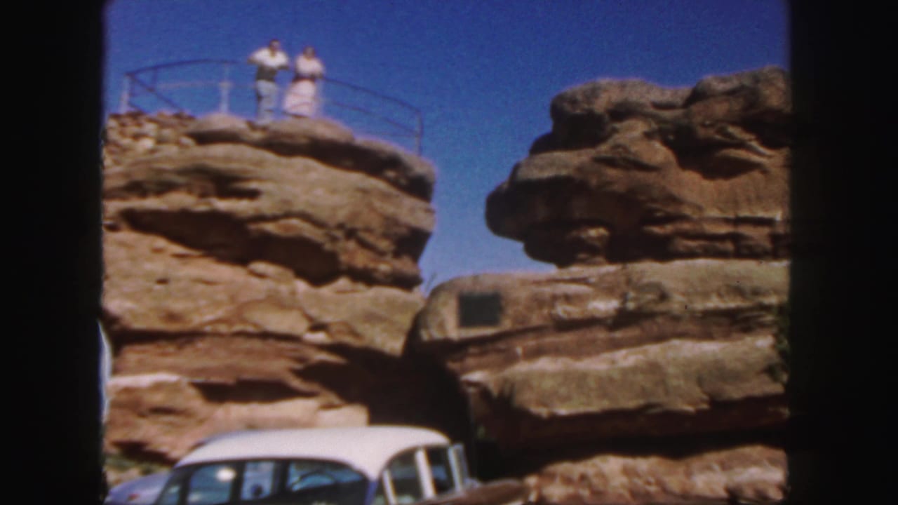 Two adults stand on a rocky overlook above a white car under a blue sky.