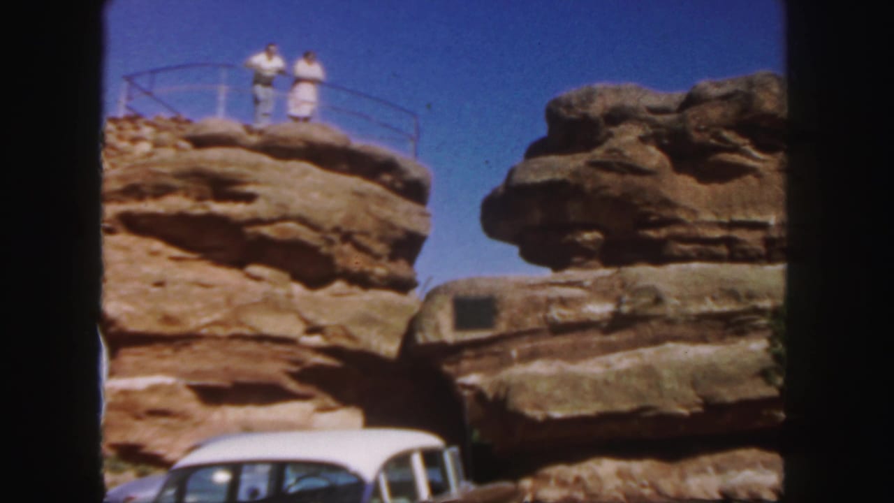 A couple stands on a Colorado rock overlook in 1958, framed by layered stone, clear sky, and a white 1950s car below.