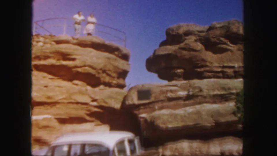 Two adults in light clothing stand on a sunlit Colorado rock overlook, with a white mid-century car cropped at the bottom.