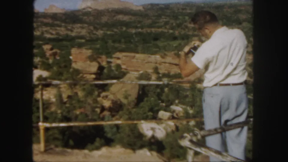 A man in 1958 Colorado stands near a railing and raises binoculars toward a canyon vista.