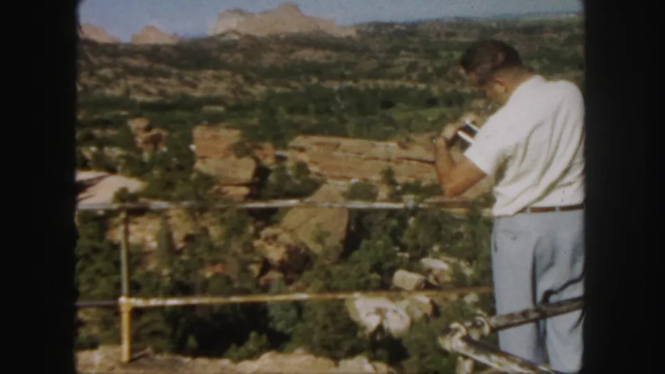 The home-movie frame shows a man with binoculars at a Colorado canyon overlook in 1958.