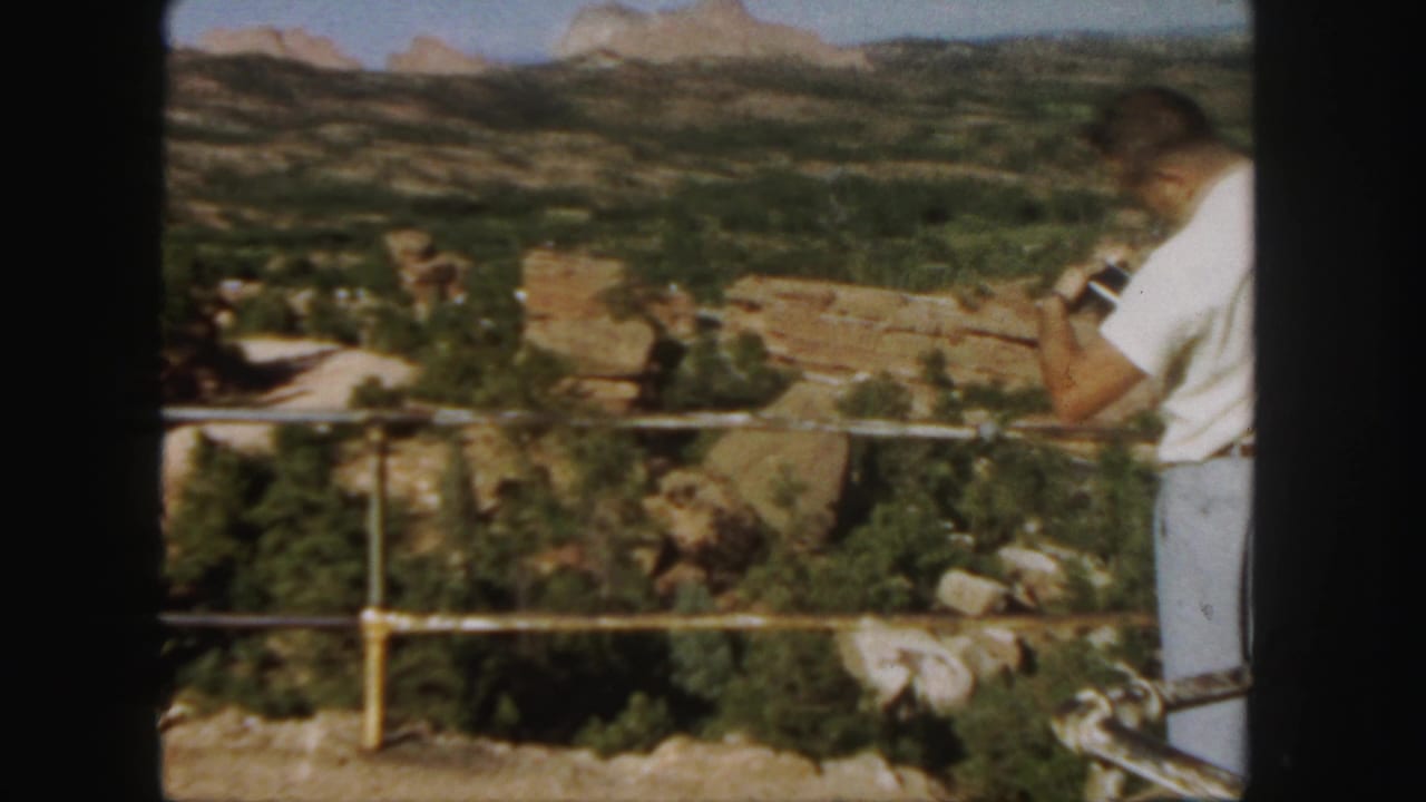 1958 Colorado home-movie frame of a man at a scenic overlook photographing a red rock canyon landscape.