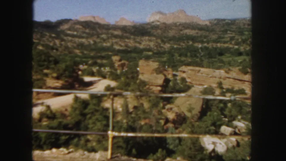 1958 Colorado 8mm view across a canyon from an overlook, with railing, red rocks, trees, and hazy mountain ridges.