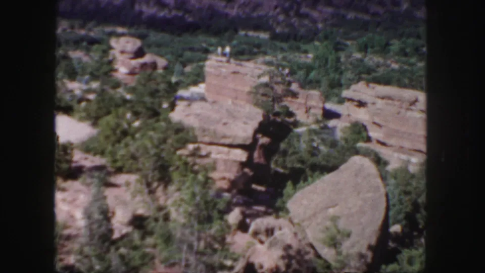 Colorado canyon home-movie frame showing scrub, boulders, red rock ledges, and visitors on a rocky outcrop in the distance.