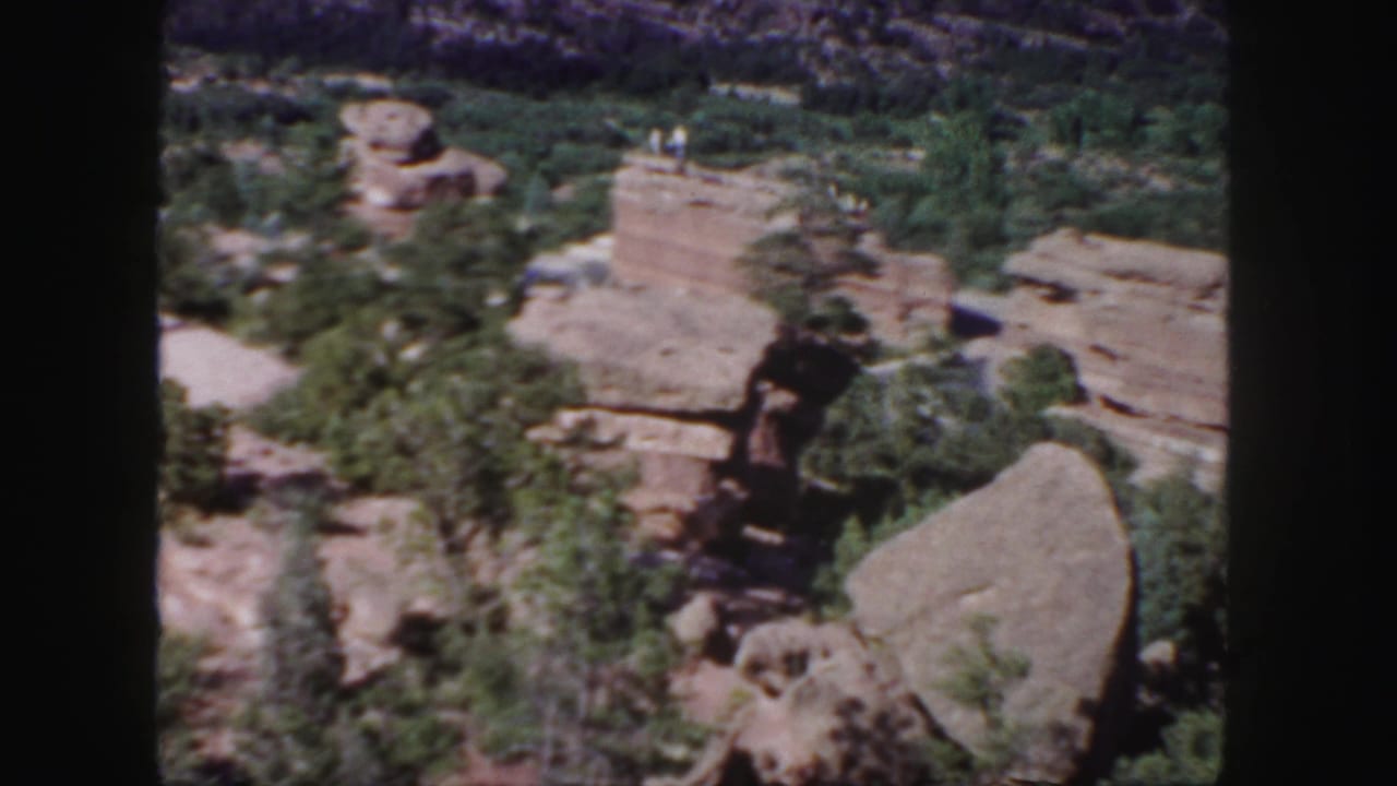 Red rock formations with scattered trees and distant people on a ledge.