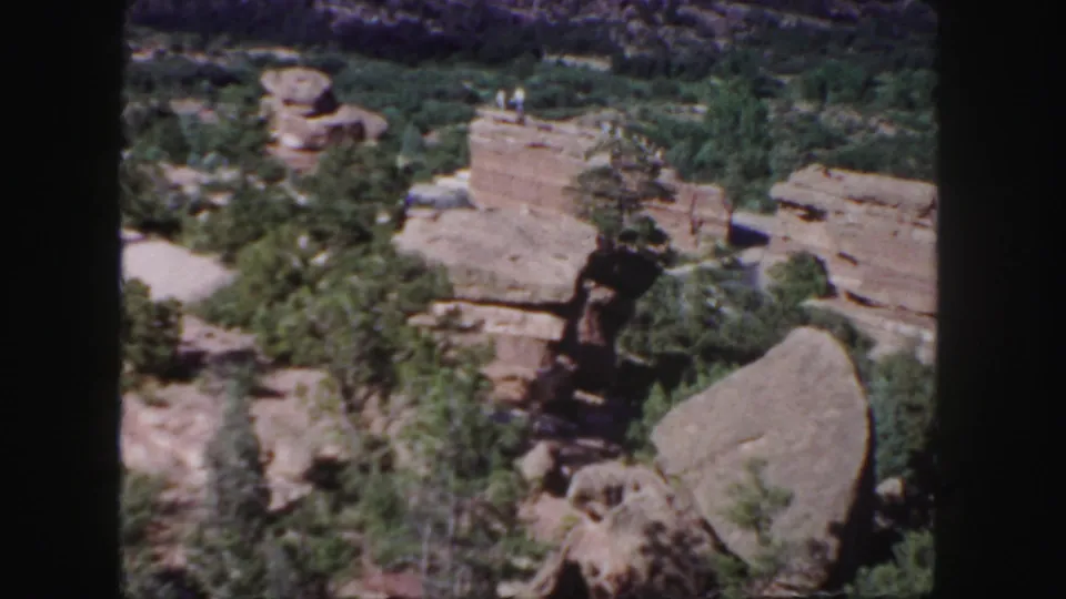 1958 Colorado landscape frame with red rock shelves, green vegetation, rounded boulders, and distant visitors on an upper ledge.