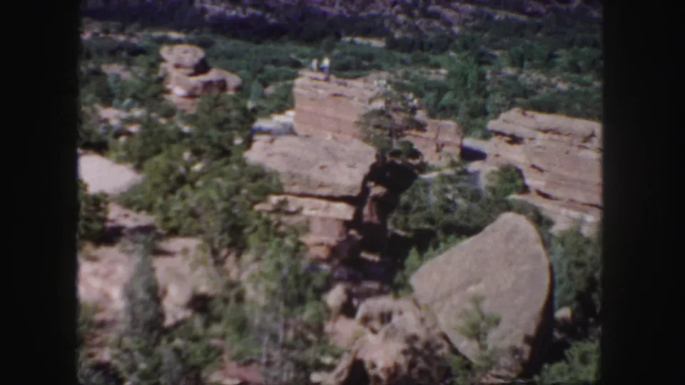 Colorado 1958 8mm frame showing a red rock outcrop, pines, scrub growth, foreground boulders, and tiny people on a high ledge.