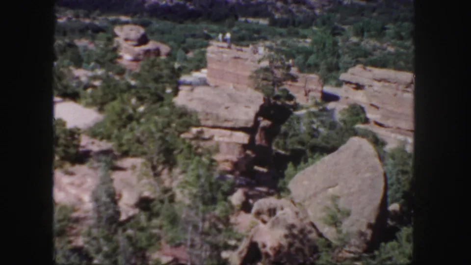 Final 1958 Colorado frame with layered sandstone, dark crevice, scattered pines, rounded boulder, and distant visitors on the upper rock ledge.