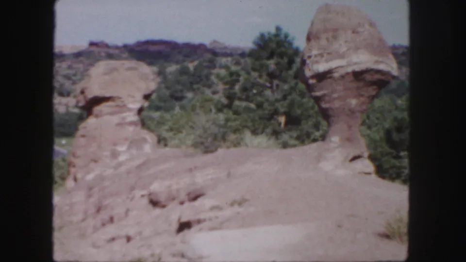 1958 Colorado 8mm frame with two weathered sandstone forms, dry terrain, green trees, and distant rocky ridges.