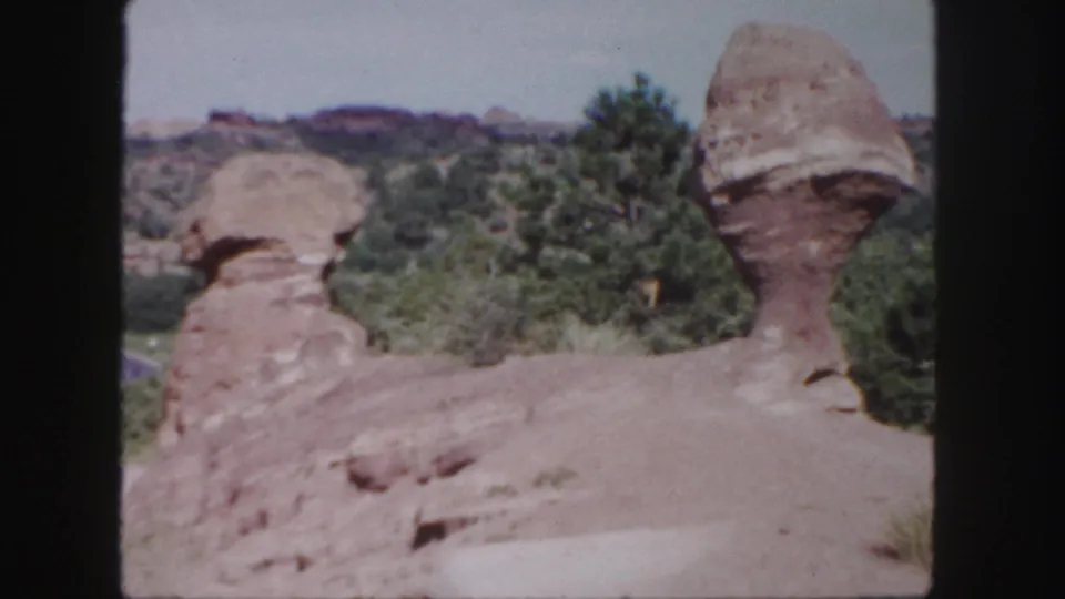 1958 Colorado home-movie frame showing rounded sandstone shapes, sparse pines, pale ground, and distant ridges.